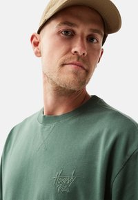 Grön sweatshirt med rund hals, prydd med texturerad sömnad och en broderad "Ärlighet Regler"-logotyp på vänster bröst.