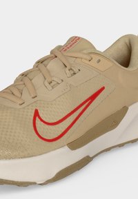 Μπεζ αθλητικό παπούτσι με κόκκινο λογότυπο Nike, επενδεδυμένο γιακά, μπεζ κορδόνια και μαλακή λευκή σόλα, σχεδιασμένο για καθημερινή ή αθλητική χρήση.