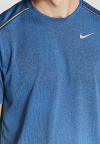 Hombre vestido con una camiseta deportiva de Nike de color azul y con un patrón, con rayas reflectantes en los hombros, mostrado desde el cuello hasta la mitad del torso.