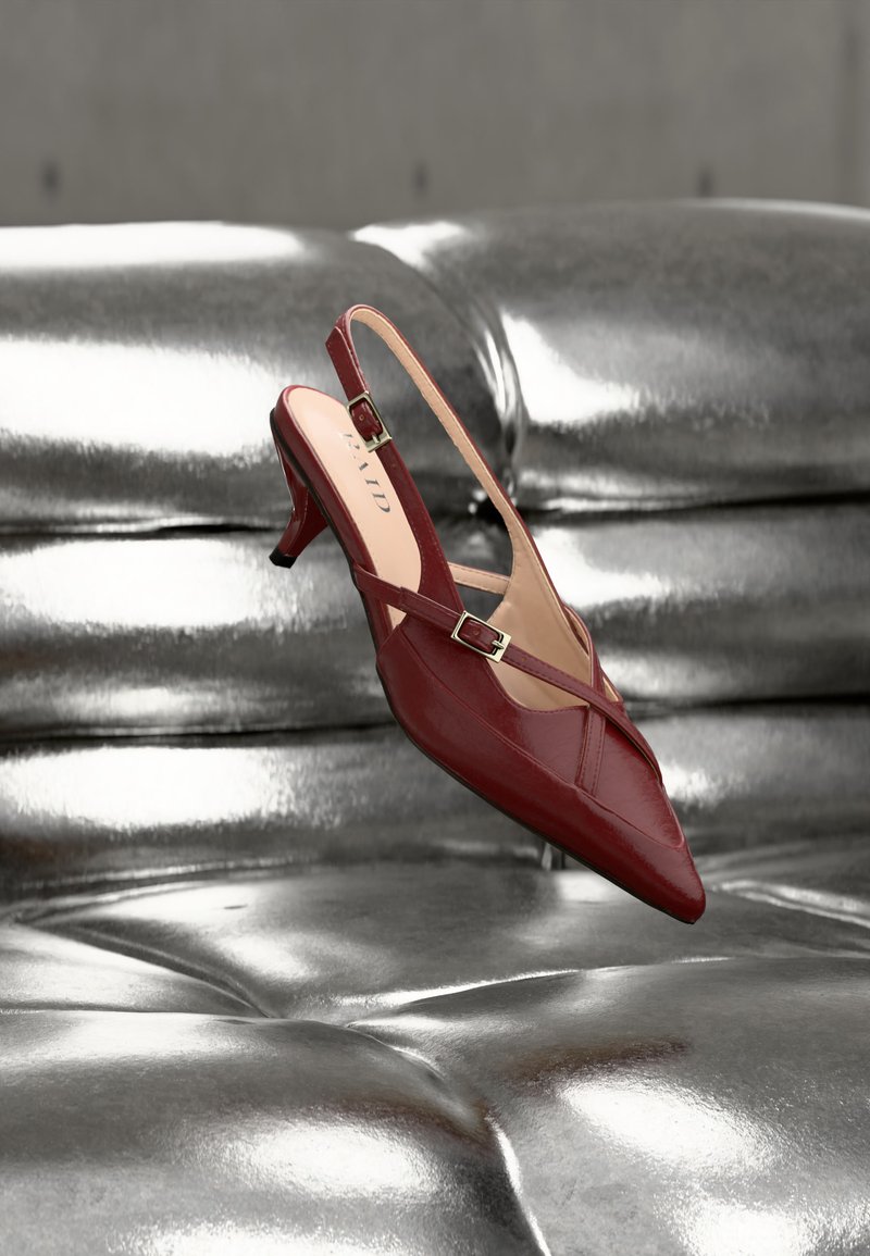 Escarpin rouge en cuir avec bride arrière, talon haut et bout pointu, doté de lanières à boucle, posé sur une surface argentée brillante capitonnée.