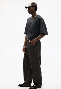 T-shirt oversize grigio scuro con il numero "89" cucito, pantaloni larghi verde oliva, collana in argento, cintura con motivo e scarpe nere.