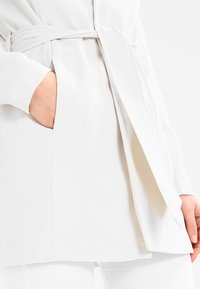 Biały, długorękawowy strój z kopertowym fasonem, teksturowaną tkaniną i wiązaniem w talii. Posiada boczną kieszeń. Gładkie, czyste linie na całej powierzchni.