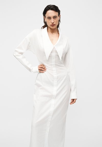 Γυναίκα ποζάρει με το χέρι στη μέση φορώντας μακρύ λευκό φόρεμα με κουμπιά, υπερμεγέθη μυτερή γιακά και μακριά μανίκια μπροστά σε απλό φόντο.