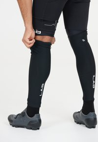 Svarta kompressionsbenärmar med en slät textur, logotyp på sidan, åtsittande design och en användare som justerar dem medan hen bär grå fotbollsskor.