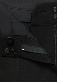 Gros plan d’un tissu noir montrant une fermeture éclair, un bouton et des crochets métalliques sur un vêtement avec des coutures détaillées.