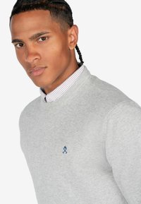 Suéter gris con cuello acanalado, que presenta un pequeño emblema azul. Debajo, se puede ver una camisa con cuello con rayas moradas y blancas.