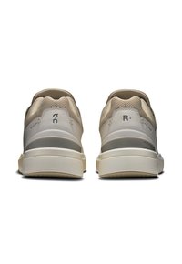 Beżowe buty sportowe z siateczkową cholewką i teksturowanym piętą. Posiadają szare akcenty i białą podeszwę. Czysty design, minimalna marka.