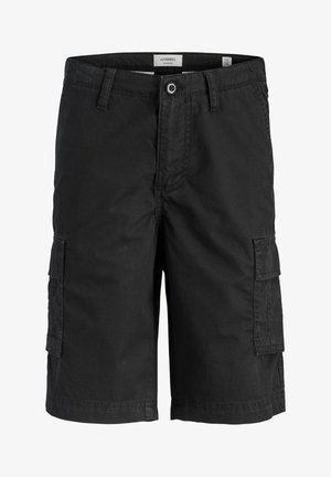 Junge steht mit einer Hand in der Tasche, trägt ein weißes T-Shirt, schwarze Cargo-Shorts, gestreifte Socken und schwarz-weiße Turnschuhe vor einem schlichten Hintergrund.