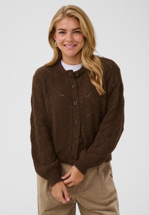 Mujer con cabello rubio largo sonriendo y vestida con un cárdigan de punto marrón abotonado y pantalones de pana beige sobre un fondo liso.