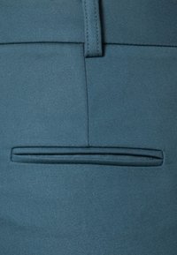 Blauwe textuurstof met een vlakke voorkant. Heeft een enkele paspelzak boven de taille. Schoon ontwerp, geen zichtbare hardware of patronen.