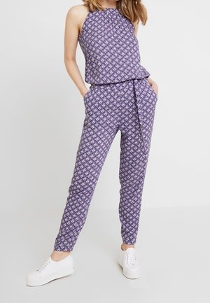 Violetti kuvioitu haalari, jossa on halterneck-kaulus, peplum-vyötärö ja kapenevat lahkeet, yhdistettynä valkoisiin lenkkareihin.