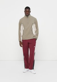 Lys grå halv-zip jakke med beige sidepaneler, parret med maroon træningsbukser med sorte sidekanter og hvidt logo, båret med hvide sko.