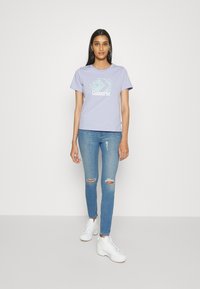 Camiseta de algodão de cor lilás clara com um logo branco "Converse", combinada com calças de ganga rasgadas de cor azul clara e sapatilhas brancas de cano alto.