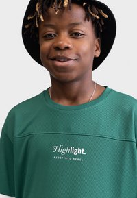 Grünes Netz-T-Shirt mit Rundhalsausschnitt und texturiertem Finish. Der Text „Highlight. REDEFINED REBEL“ ist in Weiß auf der Vorderseite aufgedruckt.