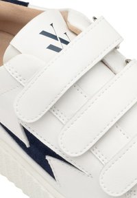 Sneaker blanche avec deux lanières auto-agrippantes, accents bleu marine et semelle blanche texturée. Design minimaliste avec un logo simple sur le talon.