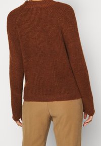 Brun stickad tröja med ribbat mönster, rundad halsringning och långa ärmar. Sömmen sitter ovanför midjan, i kombination med beiga byxor.