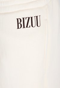Witte katoenen sweatpants met een zwart "BIZUU" logo aan de linkerkant. Ze hebben een gladde textuur en zijzakken.