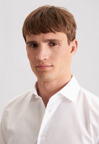 Junger Mann mit kurzem braunem Haar, der ein weißes Hemd mit Knöpfen trägt und leicht seitlich blickt, vor einem schlichten hellen Hintergrund.