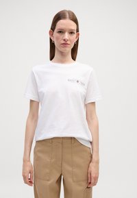 Maglietta di cotone bianca con maniche corte, con un piccolo logo grigio sul lato sinistro. Abbinata a pantaloni marrone chiaro.