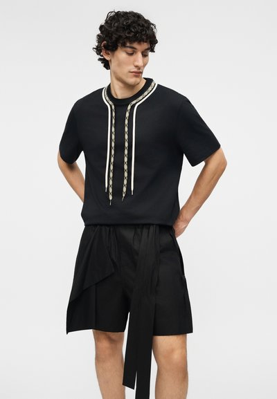 Mladý muž s kudrnatými vlasy, který má na sobě černé tričko s krátkým rukávem s vzorovanými šňůrkami a volné černé šortky s látkovými detaily.