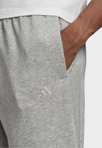 Højre hånd i lommen på lysegrå Adidas sweatpants med broderet logo, parret med en hvid top.