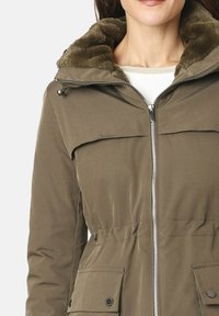 Chaqueta verde olivo con capucha forrada de piel sintética, cierre de cremallera al frente, cintura elástica y dos bolsillos con solapa; presenta una textura de tela suave.