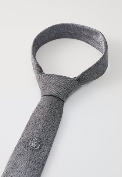 Siva pletena kravata z teksturirano površino, ki na ožjem koncu prikazuje logotip glave v krogu. Nastavljiv vratni del za boljše prileganje.