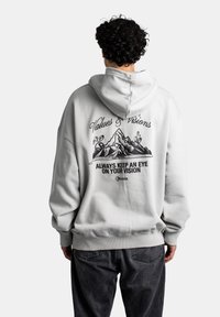Lys grå hettegenser med en frontlomme og snor i hetta. Baksiden har svart grafisk tekst og fjelldesign. Laget av bomullsblanding.