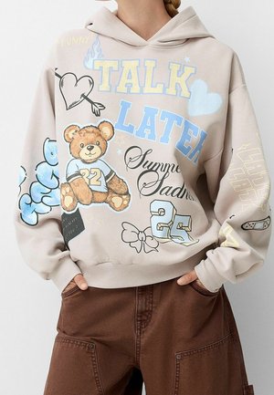 Hoodie beige avec un ours en peluche imprimé, graphiques colorés et texte. Comprend une poche kangourou et des poignets côtelés. Matériaux doux.