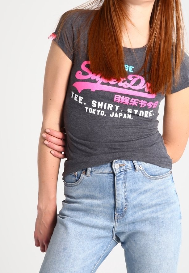 Grå t-shirt med rosa grafisk textdesign som inkluderar "SuperDry" och "Tokyo, Japan." Matchas med ljusblå slitna jeans.