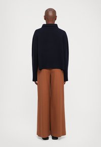 Osoba z ogolonym łbem, ubrana w ciemnoniebieski sweter i szerokie brązowe spodnie, stojąca tyłem na jednolitym białym tle.