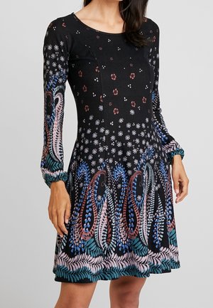 Kvinde iført en sort langærmet kjole med flerfarvede blomster- og paisleymønstre, figursyet talje og rund halsudskæring.