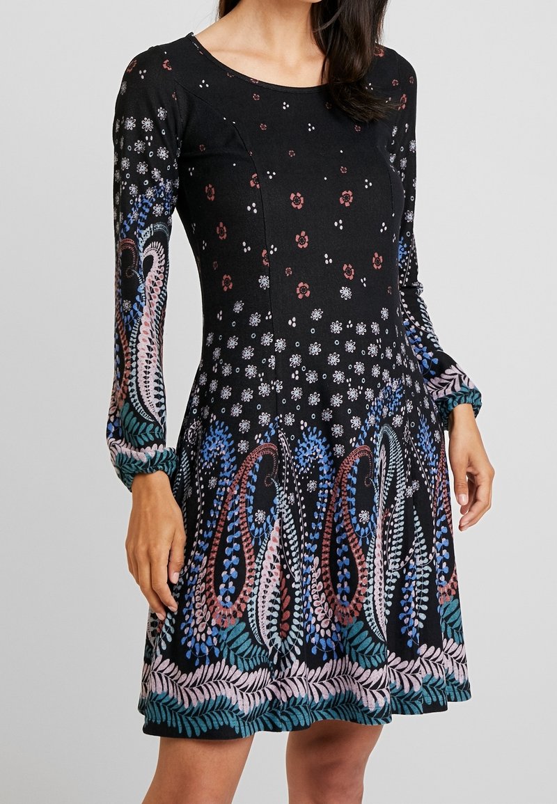Femme portant une robe noire à manches longues avec des motifs floraux multicolores et paisley, taille cintrée et encolure ronde.