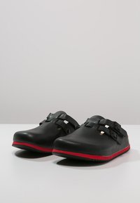 Schwarze Clogs mit glatter Textur, ausgestattet mit zwei verstellbaren Schnallen und ausgeschnittenen Details, akzentuiert durch ein rotes Band entlang der Sohle.