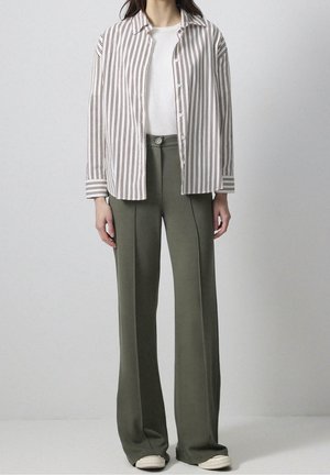 Personne portant un pantalon ample vert olive, des baskets blanches, un t-shirt blanc et une chemise blanche à boutons avec des rayures verticales marron.