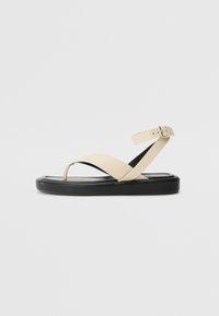 Beige leather sandals with a narrow strap design, featuring a toe post and ankle strap. The sole is thick and black with a smooth finish.