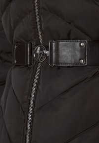 Sort quiltet pufferjakke med en metallisk lynlås, sort læderlivbånd og en cirkulær sølvspænde detalje.