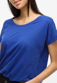 Modrá bavlněná trička s širokým kulatým výstřihem, krátkými rukávy s ohrnutím a hladkou strukturou. Detailní pohled zdůrazňující střih.