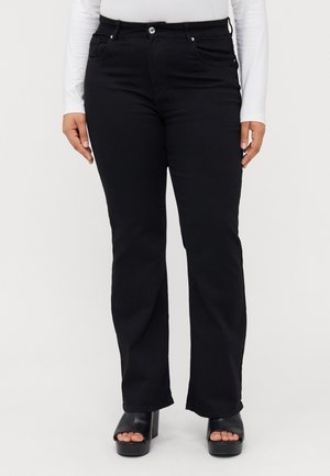 Lower body of person wearing black bootcut pants, white long-sleeve top, and black open-toe platform shoes against a plain background.