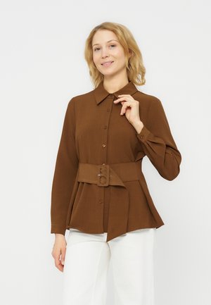 Frau mit blonden Haaren, die ein braunes, mit Gürtel versehenes Hemd mit Knöpfen und weiße Hosen trägt, posiert mit der Hand am Kragen vor weißem Hintergrund.