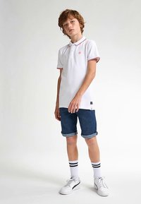 Bílá polokošile s červeným lemováním, modré džínové šortky, bílé tenisky a pruhované ponožky. Neformální design s volným střihem a detailem loga.