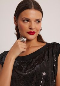 Czarny top z cekinami o wyciętym dekolcie i krótkich rękawach. Modelka nosi srebrny pierścionek oraz kolczyki z perłami, a jej usta są pomalowane na intensywny czerwony kolor.