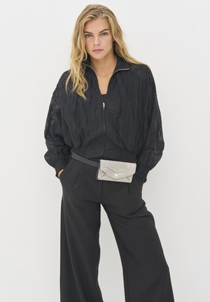 Melns krunkāts audums bomber jakas ar rāvējslēdzēju priekšpusē, apvienots ar platām melnām apakšbiksēm. Sudraba rhinestone maku atsperes uz gurniem.