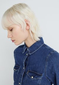 Denim shirt in deep blue with a classic collar, front buttons, and two chest pockets. Features contrasting stitching and a textured fabric.