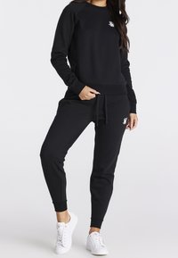 Felpa nera con polsini a costine, abbinata a pantaloni jogger coordinati. Entrambi presentano un piccolo logo bianco. Indossata con sneakers bianche.