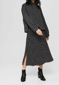 Tmavě šedá mikina s kapucí a širokými rukávy, navrstvená přes černé šaty s bílým puntíkovým vzorem a bočními rozparky, doplněná černými kotníkovými botami.