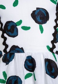 Vitt tyg med mörkblå rosor och gröna blad, detaljerat med svart scalloped kant längs sömmarna. Kjoldesign med plissering.