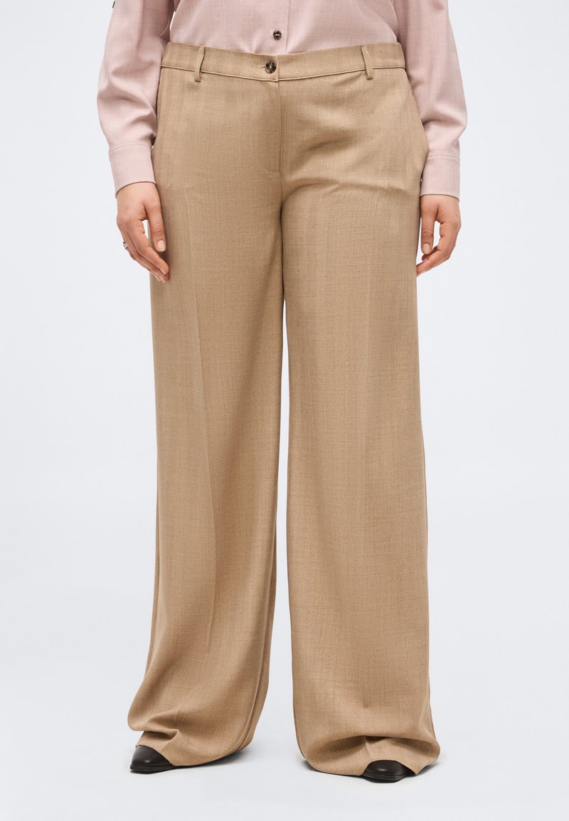 Μπεζ παντελόνια φαρδιάς γραμμής με θηλιές για ζώνη, μπροστινό κουμπί και φερμουάρ, φορεμένα με ανοιχτό ροζ μπλούζα μακρυμάνικη και μαύρα παπούτσια.