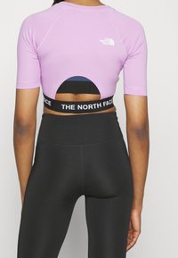 Top corto de manga corta en púrpura claro con espalda descubierta y banda elástica negra con el logo de "THE NORTH FACE", combinado con leggings negros.