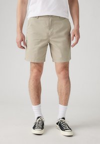 Shorts din bumbac bej cu o croială ajustată, buzunare laterale și margini răsucite, purtate cu adidași negri și șosete albe până la glezna.